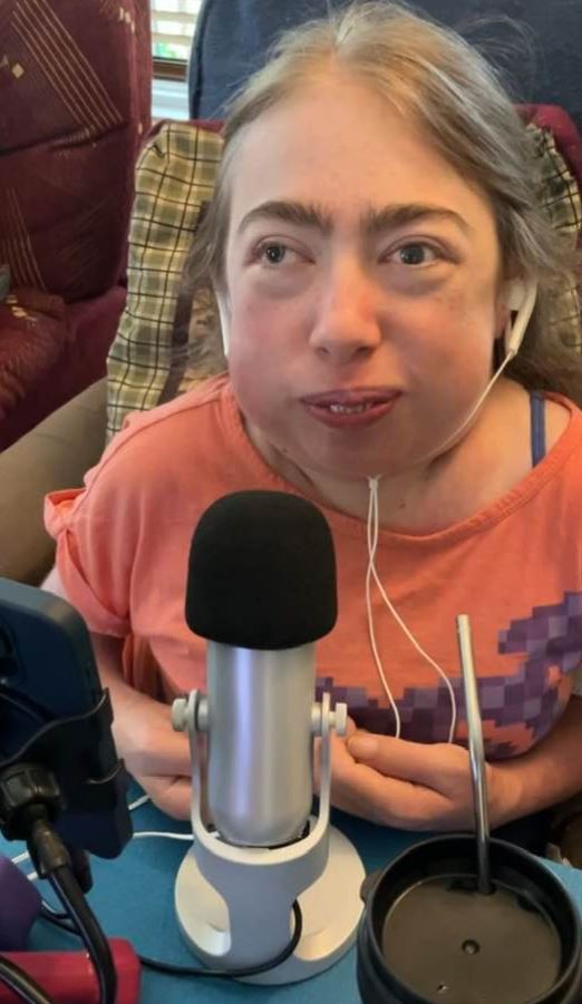 Monika sitting at a table, wearing a coral-colored shirt with a purple geometric pattern. She is equipped with bone conducting earphones, and in front of her, there is a silver microphone on a stand. Additionally, there is a smartphone mounted on a tripod positioned to her left and a black travel mug with a straw to her right on the table.